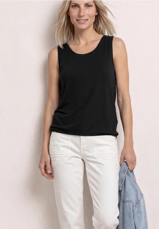 Vrouw in zwarte tanktop en witte broek, houdt een spijkerjasje vast, tegen een lichte achtergrond.