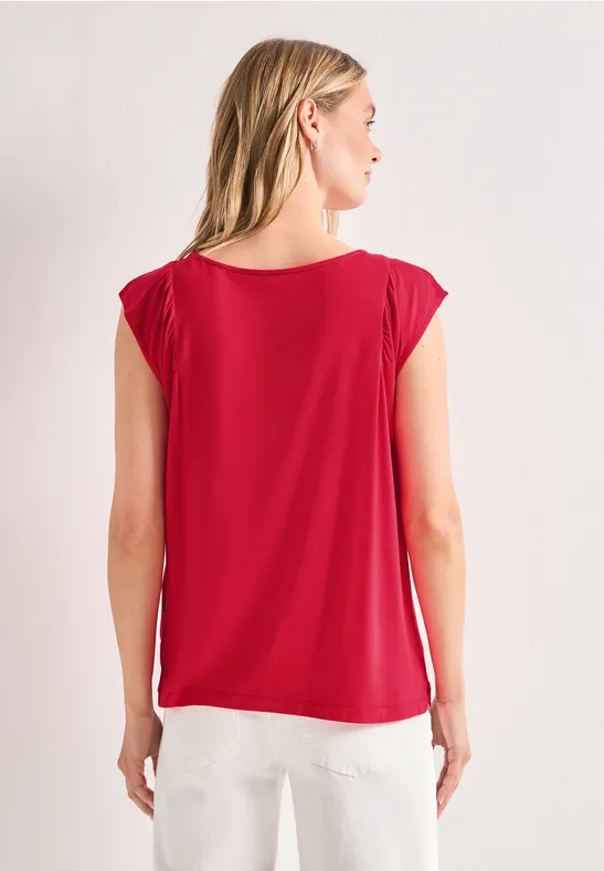 Vrouw van achteren, draagt een losse rode top met korte mouwen en een witte broek voor een neutrale achtergrond.