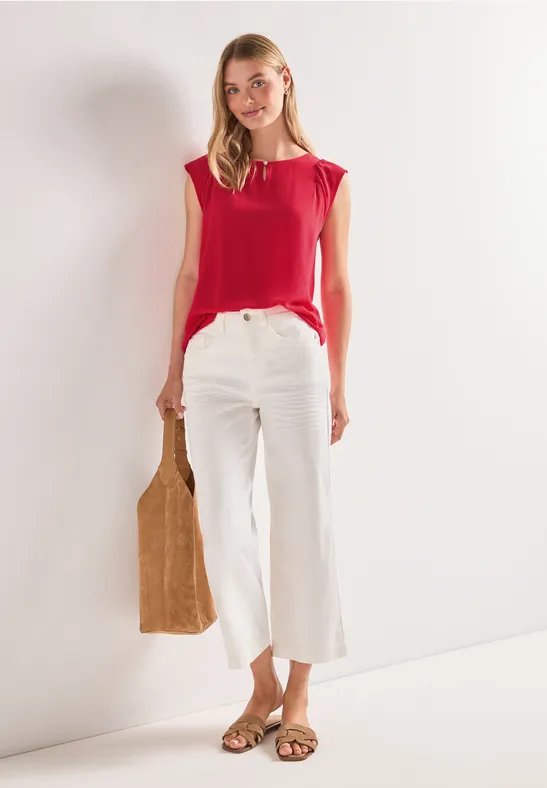 Vrouw in rood shirt en witte, wijdvallende broek, met bruine tas en platte sandalen.