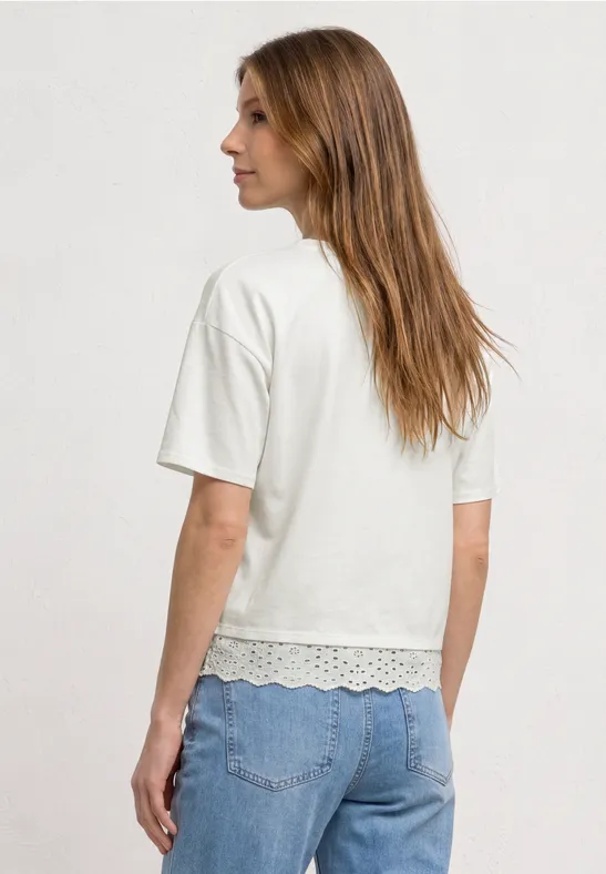 Een vrouw van achteren in een wit T-shirt met korte mouwen, met kanten zoom en blauwe spijkerbroek.