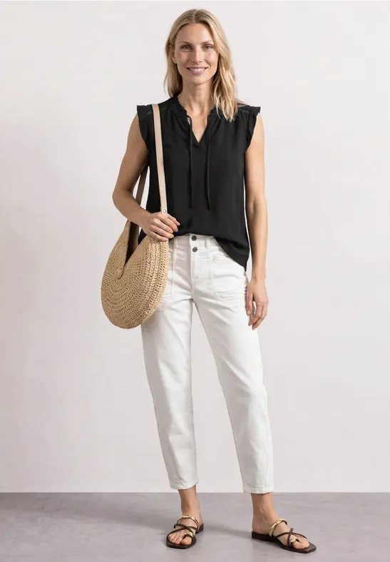 Vrouw draagt zwarte mouwloze top, witte jeans, zwarte sandalen en rieten tas.