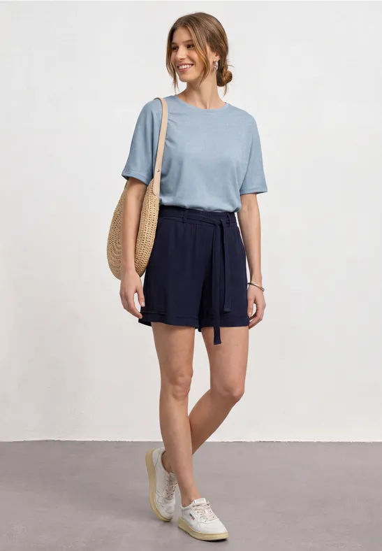Femme en t-shirt bleu, short bleu foncé, sac en paille et baskets blanches.