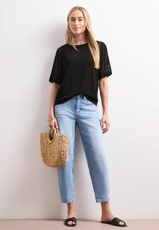 Eine schlanke Frau trägt ein schwarzes T-Shirt, helle Jeans, schwarze Sandalen und hält eine Basttasche.