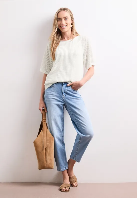 Frau in weißem Oberteil und hellblauen Jeans, trägt eine Tasche und Sandalen, vor weißem Hintergrund.