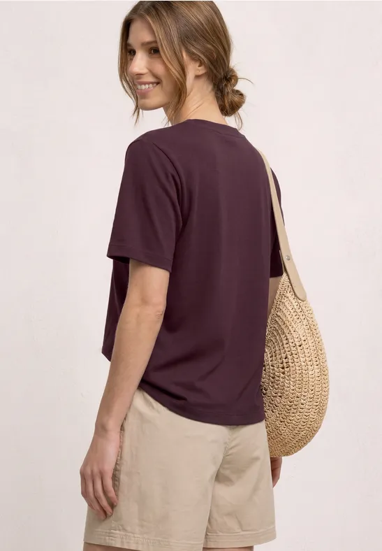 Vrouw gezien van achteren in paars T-shirt, beige shorts en een ronde stro-tas.