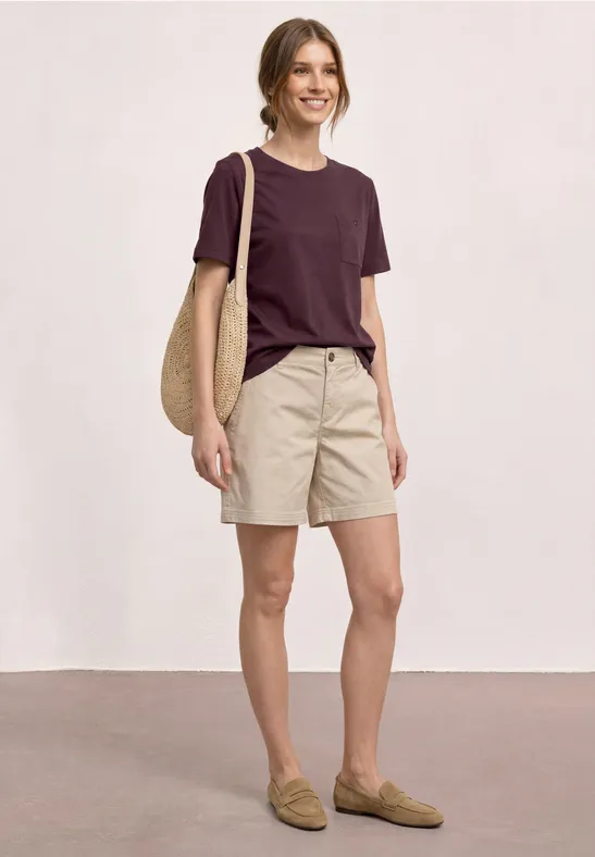 Persoon in casual kleding: bordeaux t-shirt, beige short, loafers en strooien tas.