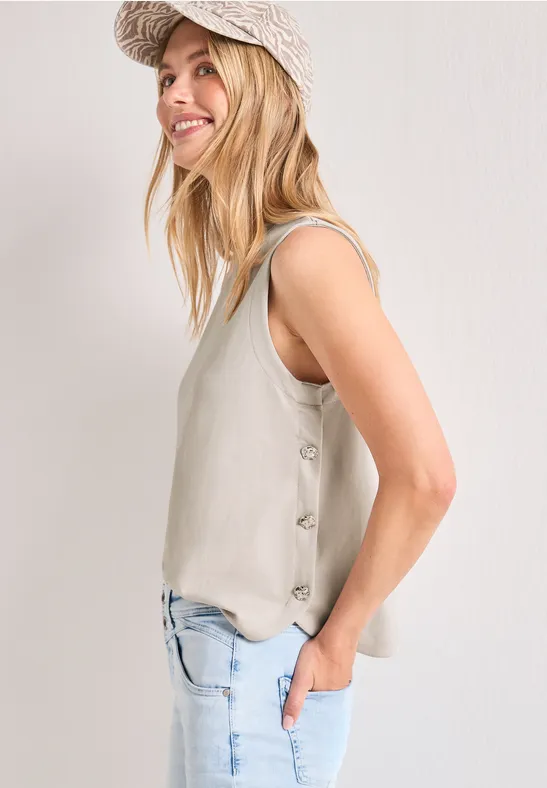 Blondine in zijaanzicht, draagt een beige mouwloze top met knopen, lichtblauwe jeans en een beige pet.