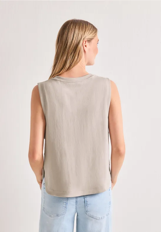 Vrouw van achteren in een beige mouwloos shirt en lichte spijkerbroek, handen in haar zakken.