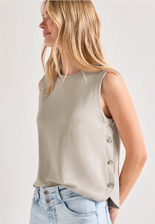 Vrouw in mouwloze beige top met zijknopen en een lichte spijkerbroek, opzij staand.