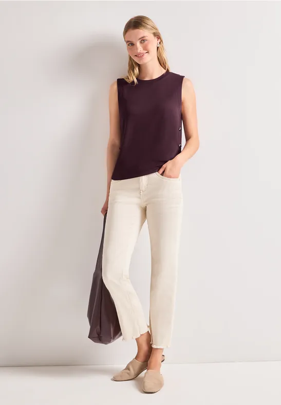 Vrouw in paarse mouwloze top en crèmekleurige broek, met bijpassende tas en instappers.