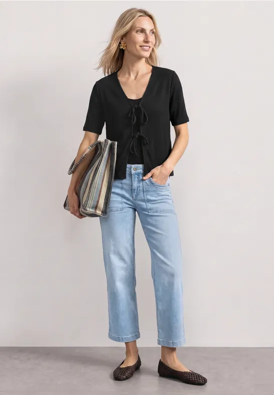 Vrouw in zwart t-shirt met korte mouwen, lichtblauwe jeans, bruine ballerina’s en een gestreepte tas voor een neutrale achtergrond.
