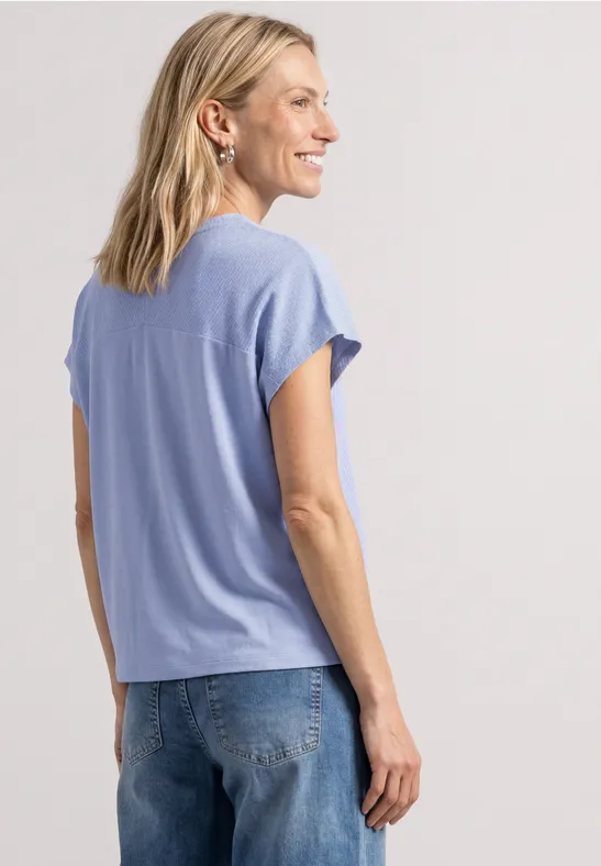 Une femme aux cheveux blonds porte un t-shirt bleu clair et un jean, de profil, sur fond neutre.