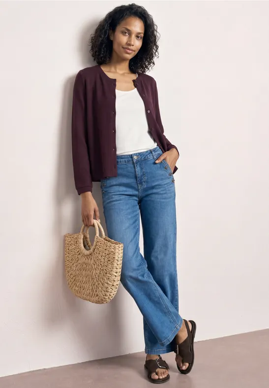 Een persoon draagt een spijkerbroek, een wit topje, een bordeaux cardigan, zwarte sandalen en houdt een strooien tas.