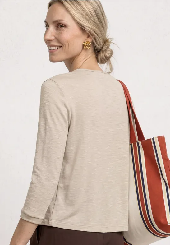 Femme vue de dos, cheveux blonds attachés, t-shirt beige 3/4 et sac rayé rouge et blanc.