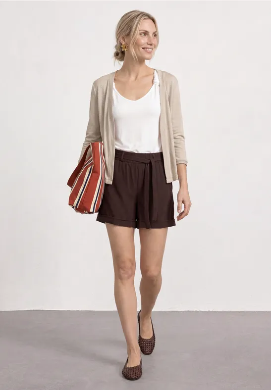 Femme en t-shirt blanc, gilet beige, short marron et mocassins, portant un sac bandoulière rayé.