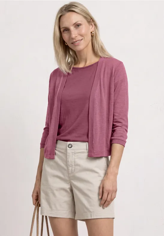 Blonde vrouw in roze shirt en vest met beige short en bijpassende tas voor een witte achtergrond.