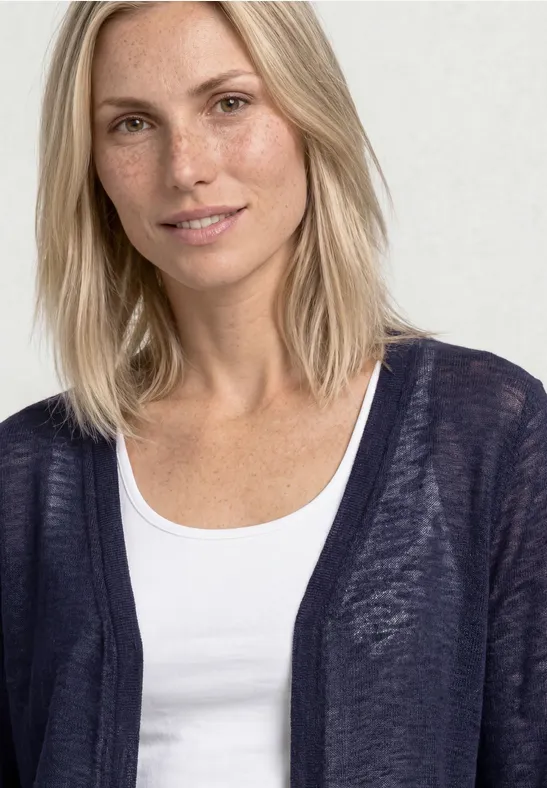 Une femme aux cheveux blonds mi-longs porte un débardeur blanc et un gilet bleu foncé.