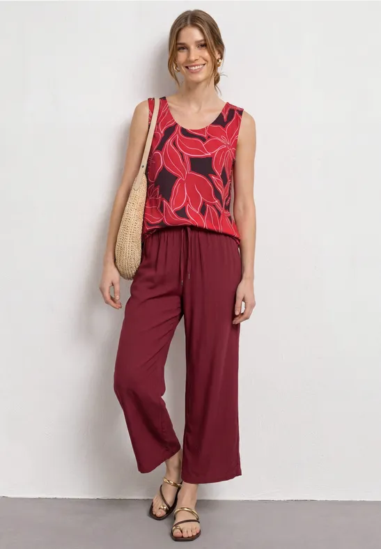 Vrouw in mouwloze top met rode bladprint, wijde bordeauxrode broek, sandalen en gevlochten tas.
