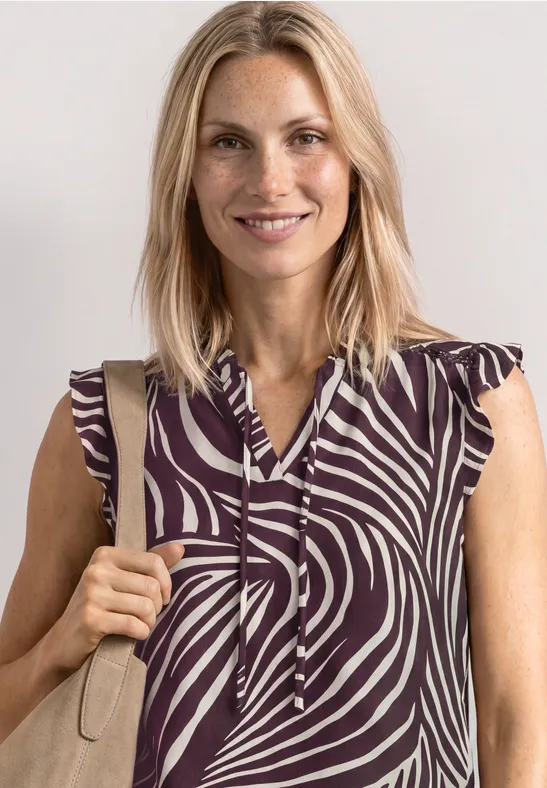 Blonde vrouw draagt een mouwloze top met paars-beige zebraprint en houdt een beige tasriem vast.