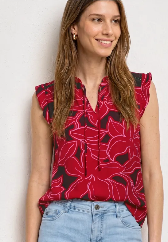 Vrouw draagt rode mouwloze top met bloemenprint en blauwe jeans voor een lichte achtergrond.