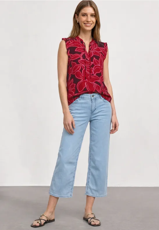 Model draagt rode mouwloze top met bloemenprint, lichtblauwe wijde jeans en zwarte sandalen voor witte achtergrond.