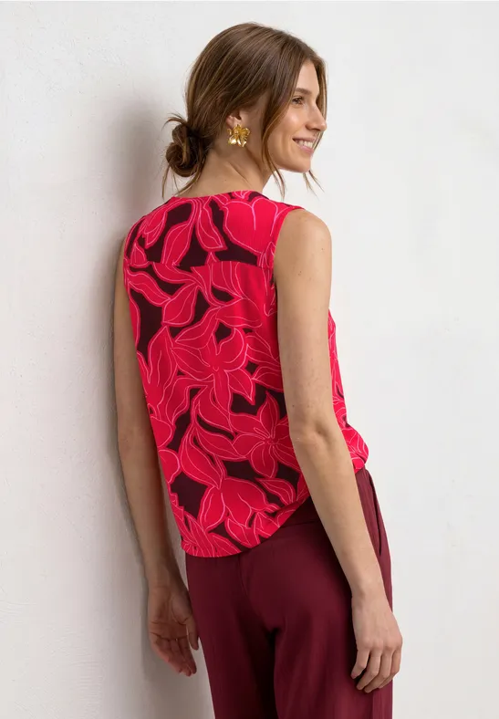 Rugaanzicht van vrouw met opgestoken haar, rood blousje met bladprint en bordeauxrode broek op lichte achtergrond.