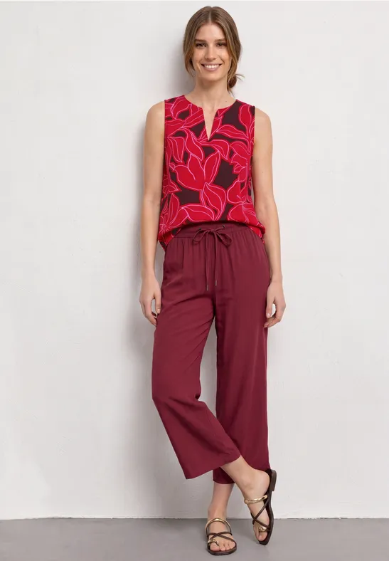 Vrouw staat voor witte muur in rood topje met bladmotief, wijde bordeauxrode broek en zwarte sandaaltjes.