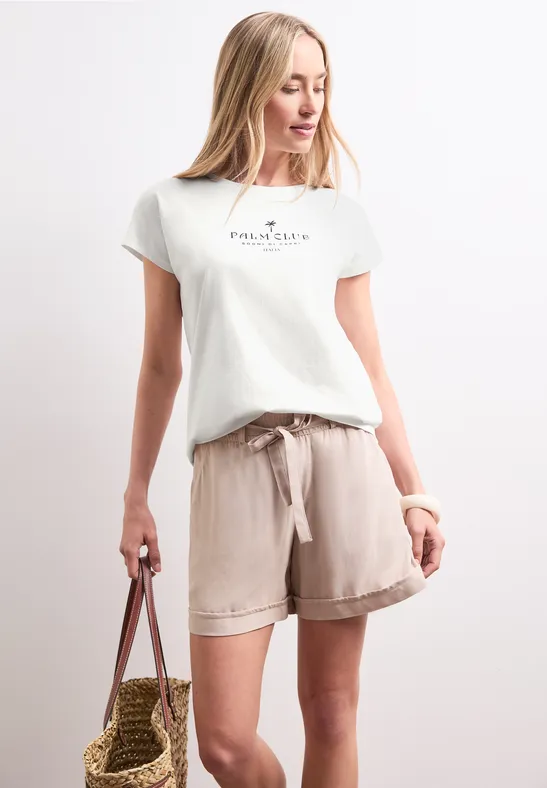 Frau in weißem T-Shirt mit Aufdruck, beigen Leinen-Shorts und Strohtasche vor weißem Hintergrund.