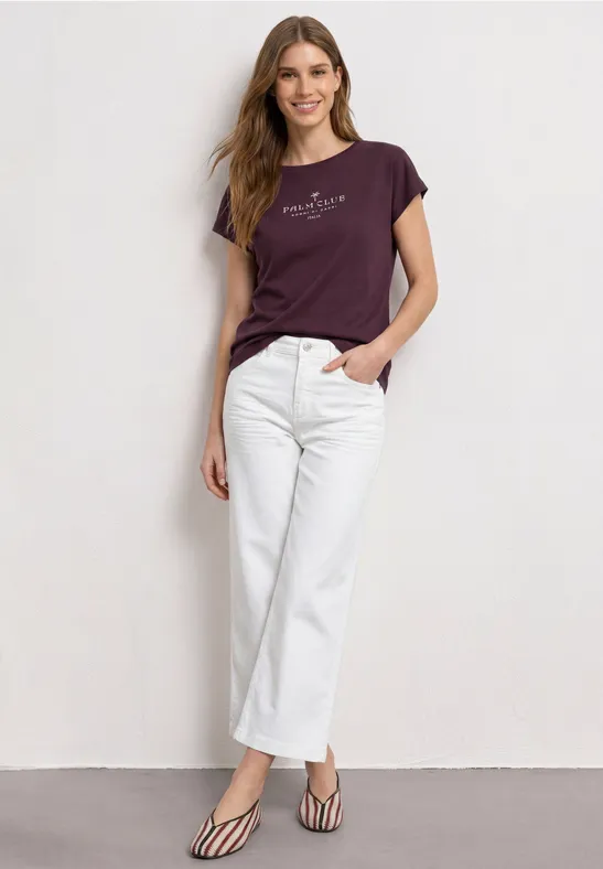 Een vrouw staat voor een muur in witte jeans, paars T-shirt en gestreepte pumps.
