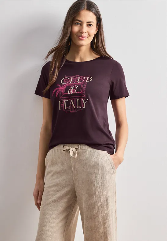 Vrouw in casual outfit: bordeaux T-shirt met 'Club Italy' en lichte linnen broek.