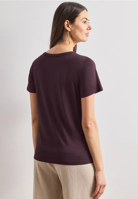 Vrouw van achteren in een donkerpaars T-shirt en lichtbeige broek tegen een witte achtergrond.