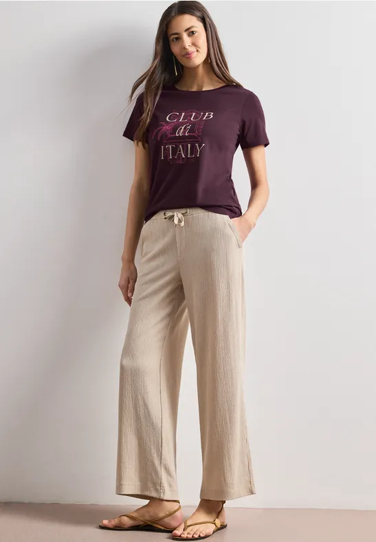 Een vrouw poseert in een minimalistische setting, draagt een paars 'Club de Italy' shirt, beige wijde broek en sandalen.