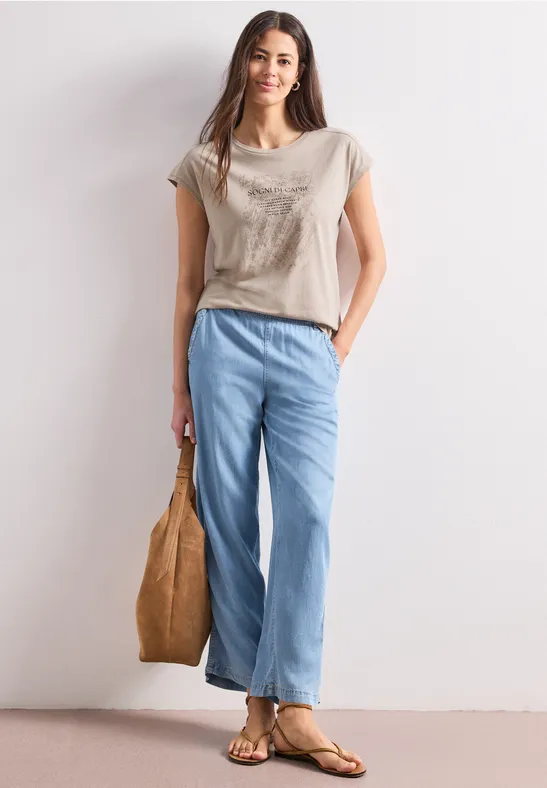 Vrouw in casual outfit: beige T-shirt, wijde blauwe jeans, sandalen en een handtas.