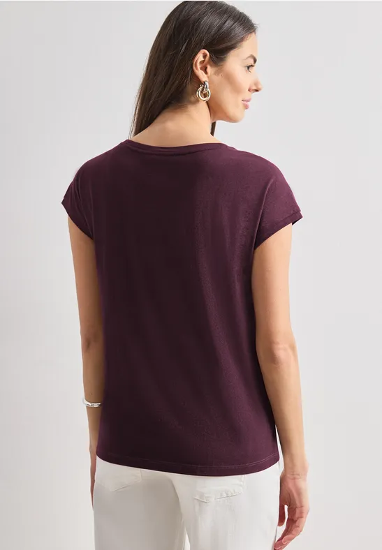Vrouw van achteren in een paars T-shirt en witte broek, lang haar, oorbellen en armband.