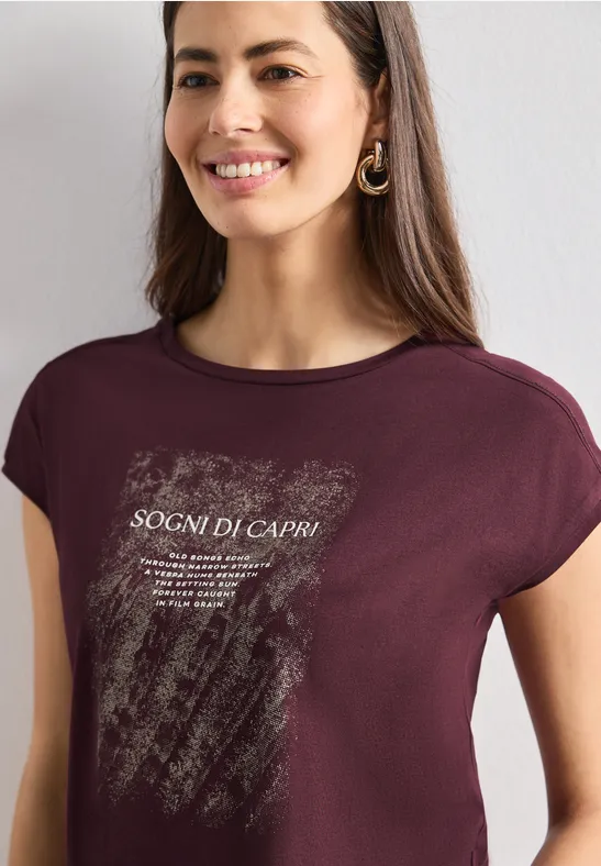 Vrouw draagt bordeauxrode t-shirt met witte print en tekst „SOGNI DI CAPRI“, glimlachend.