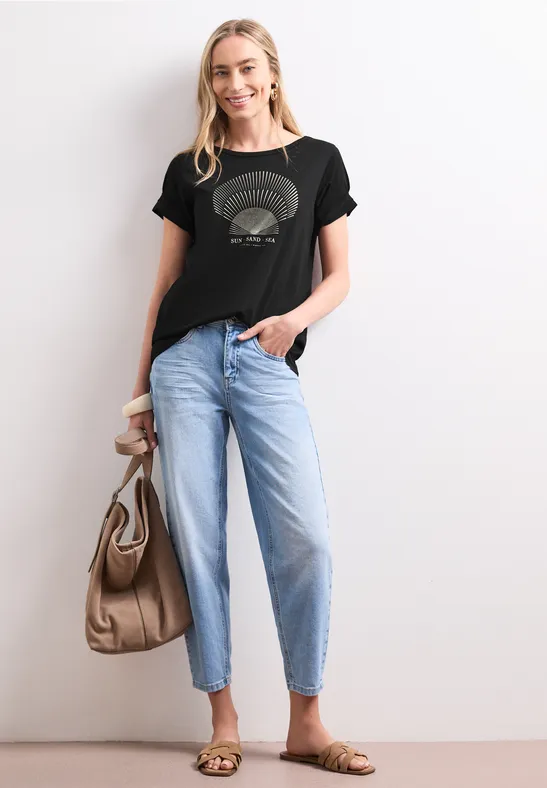 Frau in lässigem Outfit: schwarzes T-Shirt mit Print, helle Jeans, Sandalen und Handtasche.