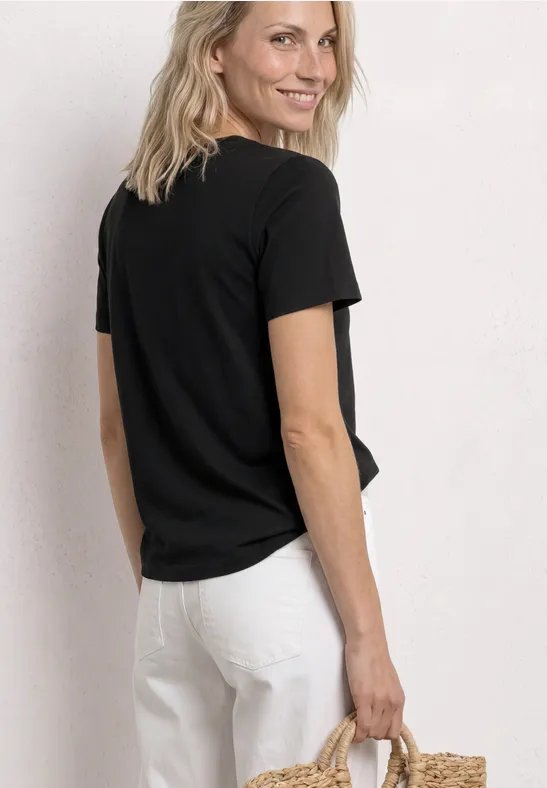 Person von hinten in schwarzem T-Shirt und weißer Hose, hält eine Strohtasche.