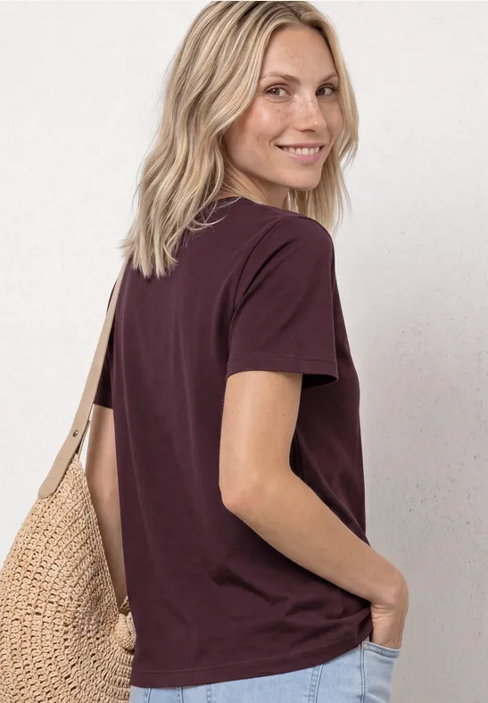Vrouw met blond haar, van achter gezien, draagt een donkerpaars T-shirt en een rieten tas.