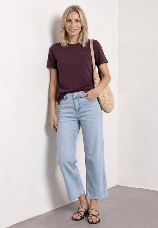 Vrouw leunt tegen witte muur, paars T-shirt, lichte jeans, zwarte sandalen en rieten tas.