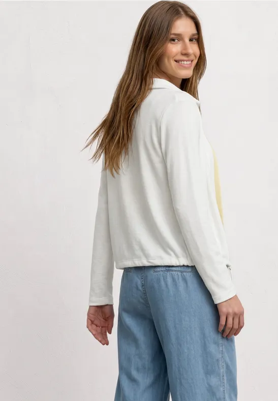 Frau von der Seite in weißer, leichter Jacke mit langen Ärmeln und blauen Jeans.