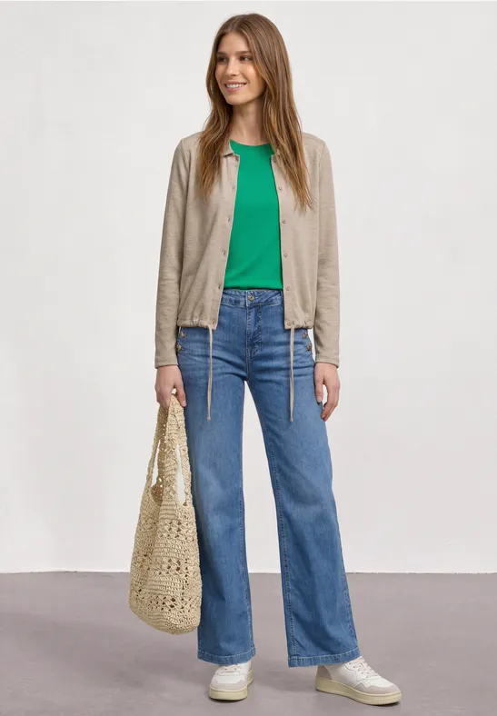 Jongevrouw met groen shirt, beige jasje, blauwe jeans, witte sneakers en een gehaakte tas.