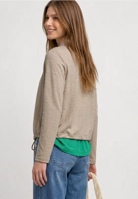 Vrouw van achteren in beige longsleeve, groen topje en spijkerbroek met een schoudertas.