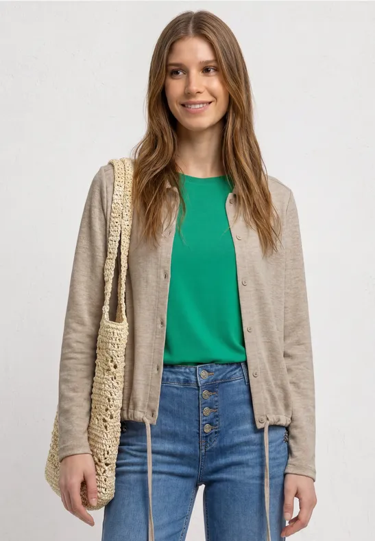 Persoon draagt beige vest, groen shirt, spijkerbroek en rieten tas.