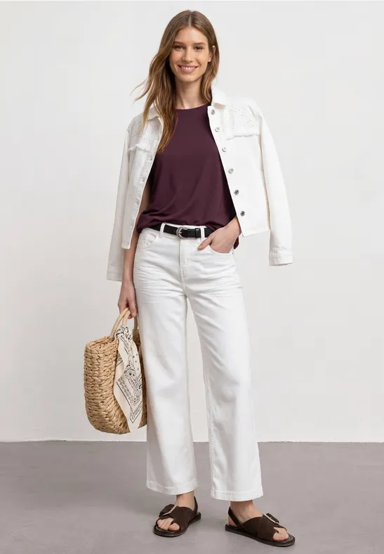 Iemand draagt een witte spijkerjas en broek, een donker shirt, een strooien tas en zwarte sandalen.