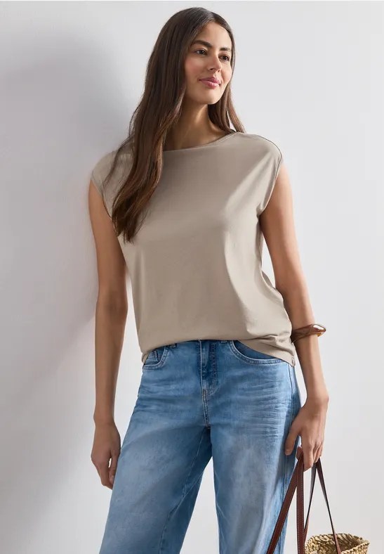Vrouw in beige mouwloos topje en blauwe jeans, met een bruine tas in de hand.