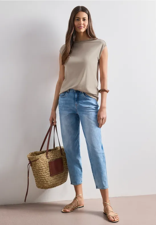 Vrouw met beige top, lichtblauwe jeans, sandalen en grote rieten tas voor een neutrale achtergrond.