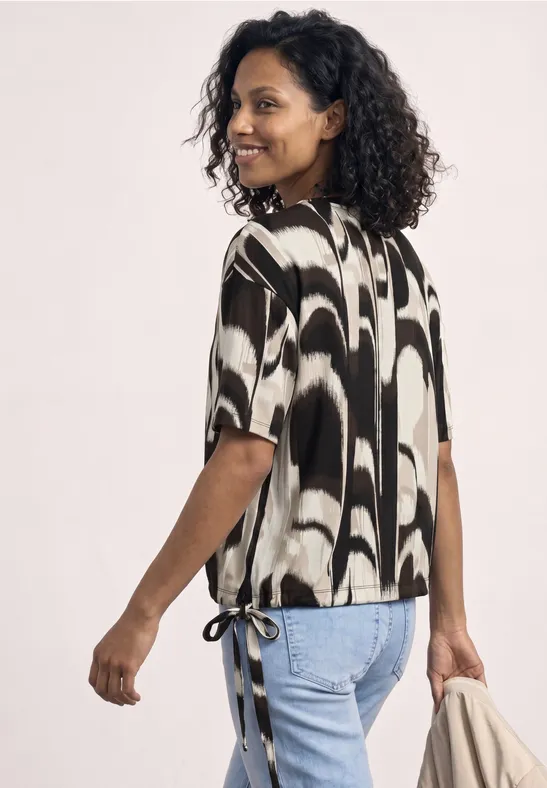 Vrouw met krullend haar van achteren, draagt een zwart-wit top met zijknopen, spijkerbroek en tas.
