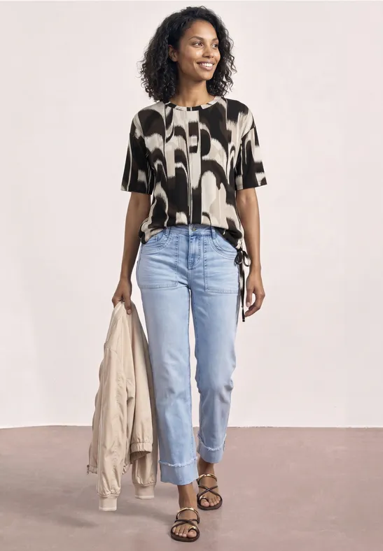 Vrouw in printblouse, lichtblauwe jeans en sandalen, met een beige jas in haar hand.