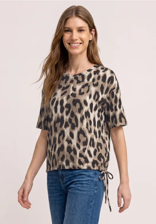 Vrouw draagt een top met korte mouwen in luipaardprint, zijdelingse tunnelkoord en blauwe jeans.