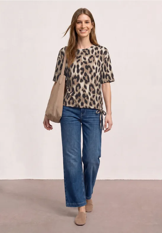 Een vrouw draagt een leopardprintshirt, blauwe jeans, beige loafers en heeft een beige tas voor neutrale achtergrond.